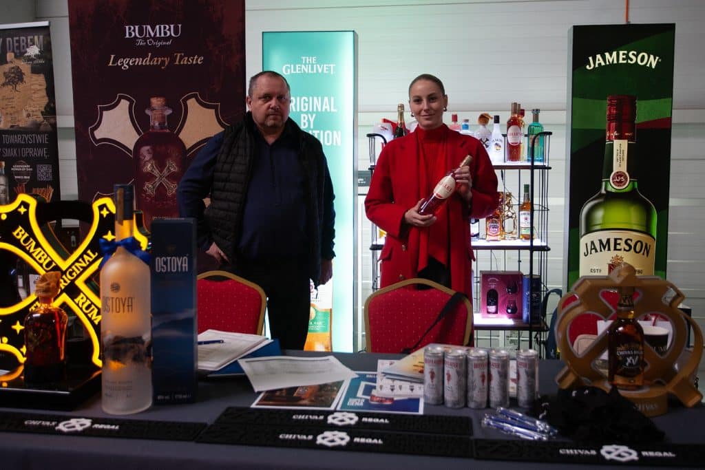 Uczestnicy targ&oacute;w w Gdańsku prezentują produkty alkoholowe na stoisku, w tle reklamy i p&oacute;łki z napojami.