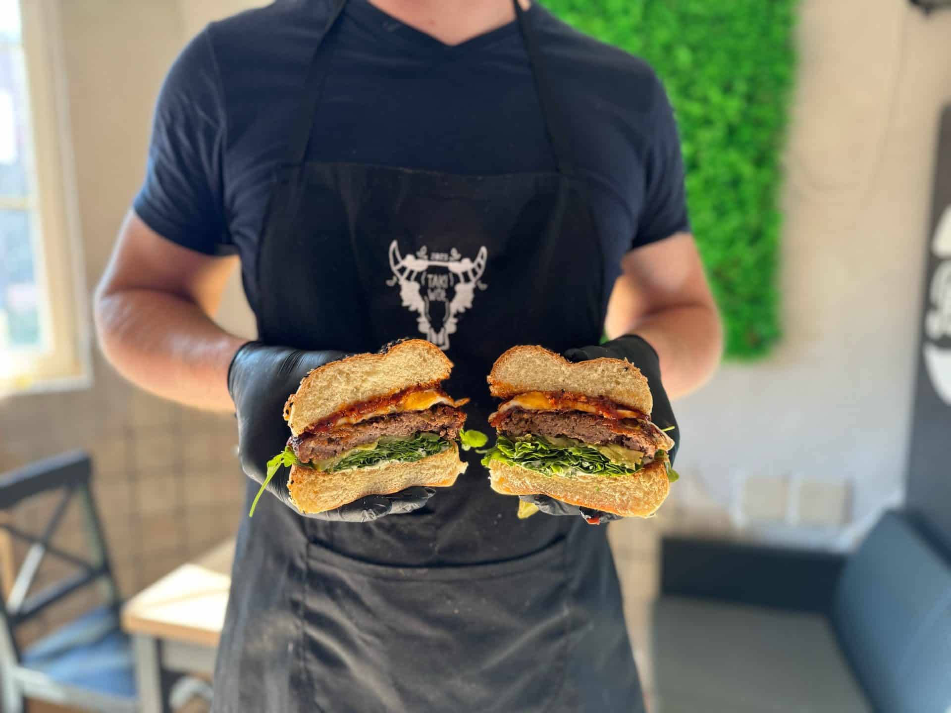 Osoba trzymająca przekrojony burger z mięsem, warzywami i serem w restauracji.