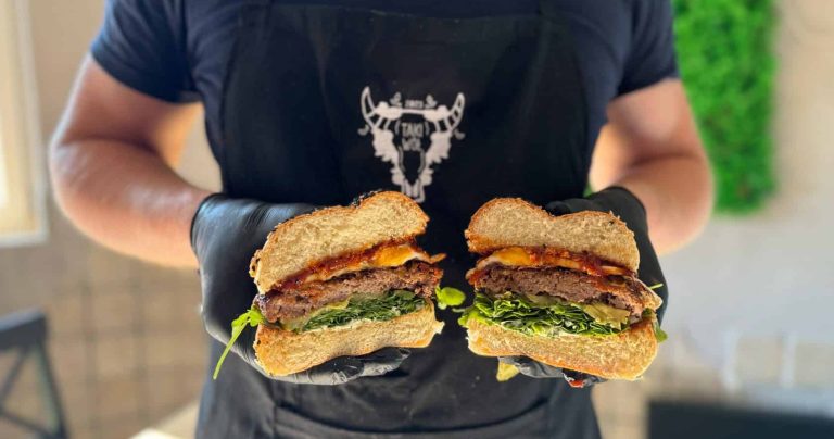 Osoba trzymająca przekrojony burger z mięsem, warzywami i serem w restauracji.