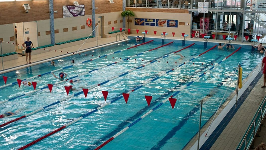 Basen pływacki w Iławie z kilkoma osobami pływającymi, widok na tor i obsługę przy krawędzi.