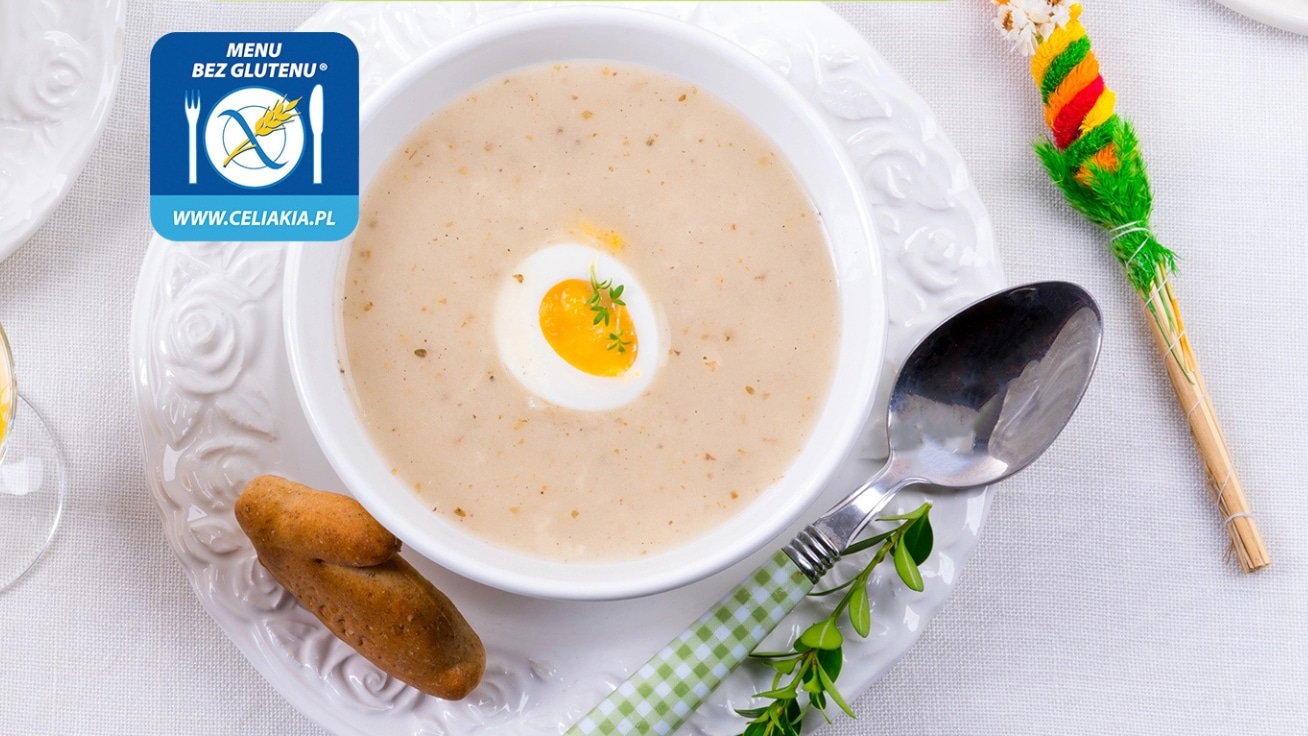 Zupa w białej misce z jajkiem na twardo, obok łyżka i chlebek, na świątecznym stole.