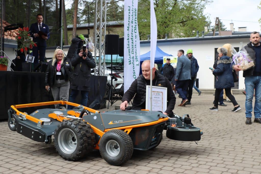 Mężczyzna z nagrodą przy dużym robotycznym urządzeniu na targach ogrodniczych w Olsztynie.