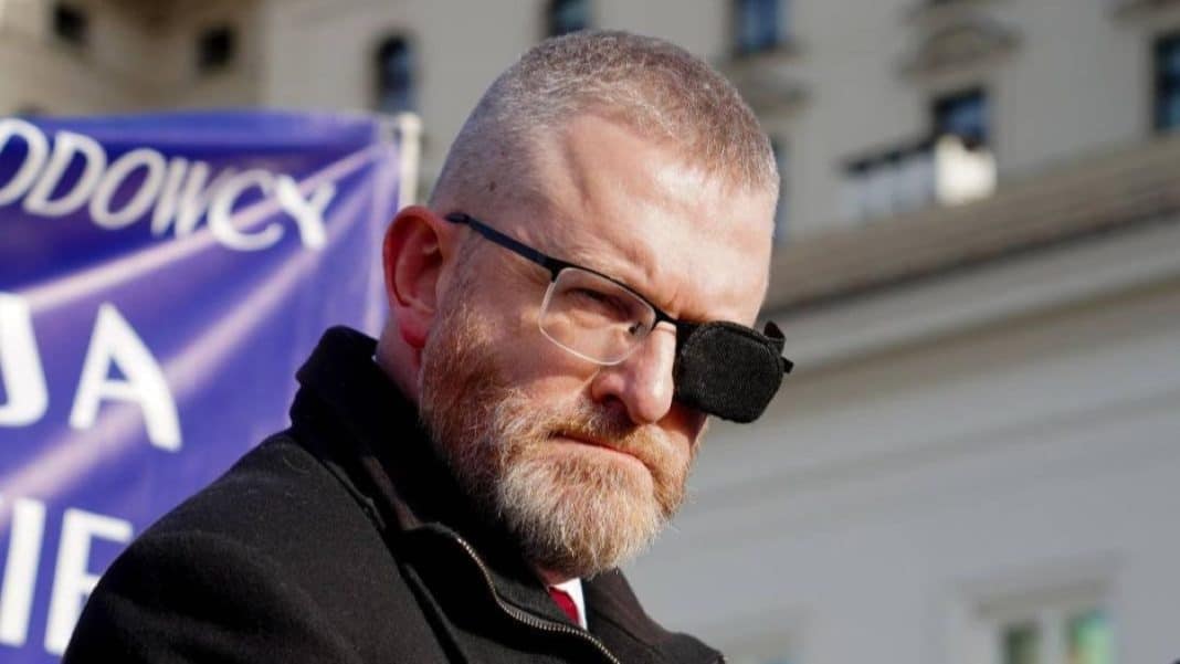 Mężczyzna z brodą i okularami, na jednym oku ma opaskę, na tle budynk&oacute;w i flagi.