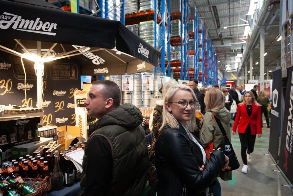 Tłum uczestnik&oacute;w na targach w hali magazynowej, stoiska z produktami i branżowe rozmowy.