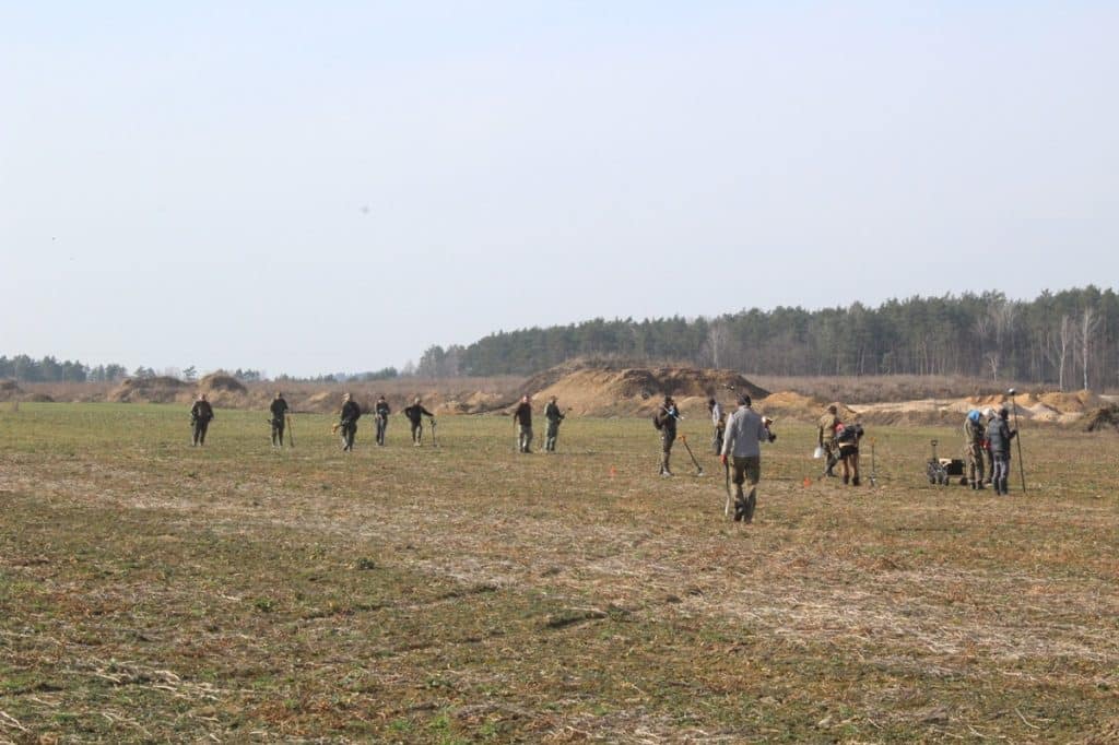 Grupa archeolog&oacute;w na polu podczas wykopalisk, w tle widoczne wykopaliska i las.