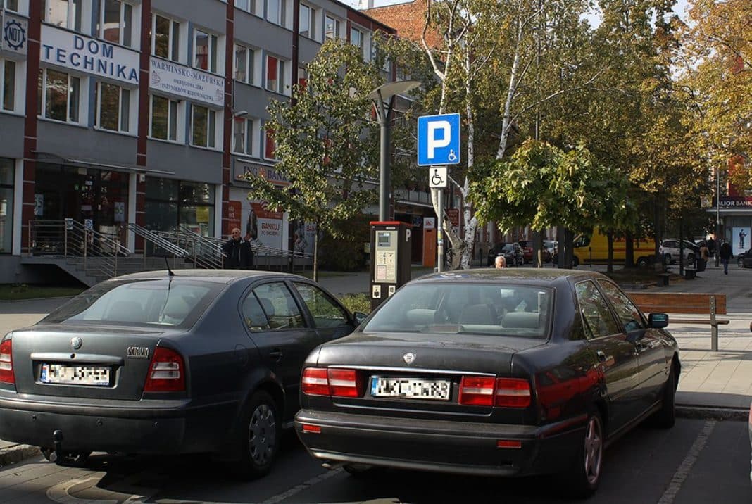 Dwa samochody zaparkowane przy parkingu z oznaczeniem dla os&oacute;b niepełnosprawnych w centrum miasta, w tle budynek z drzewami.