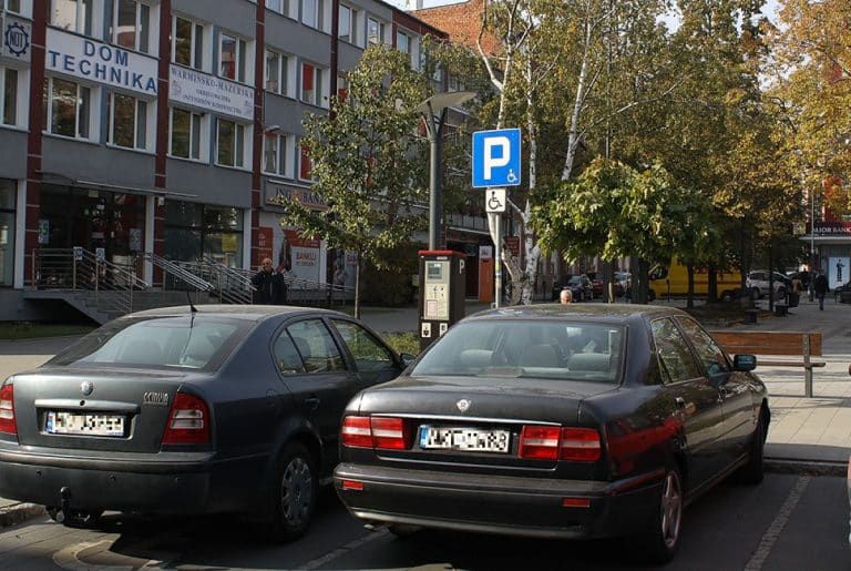Dwa samochody zaparkowane przy parkingu z oznaczeniem dla os&oacute;b niepełnosprawnych w centrum miasta, w tle budynek z drzewami.