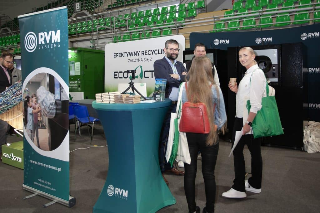 Uczestnicy rozmawiają przy stoisku na targach w hali sportowej, promując recykling i ekologiczne rozwiązania.