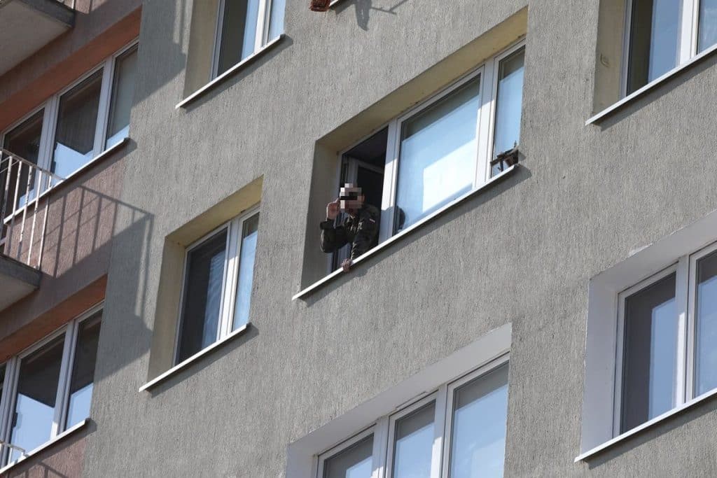 Mężczyzna rozmawia przez telefon z okna mieszkania na wysokim piętrze budynku, wok&oacute;ł służby ratunkowe.