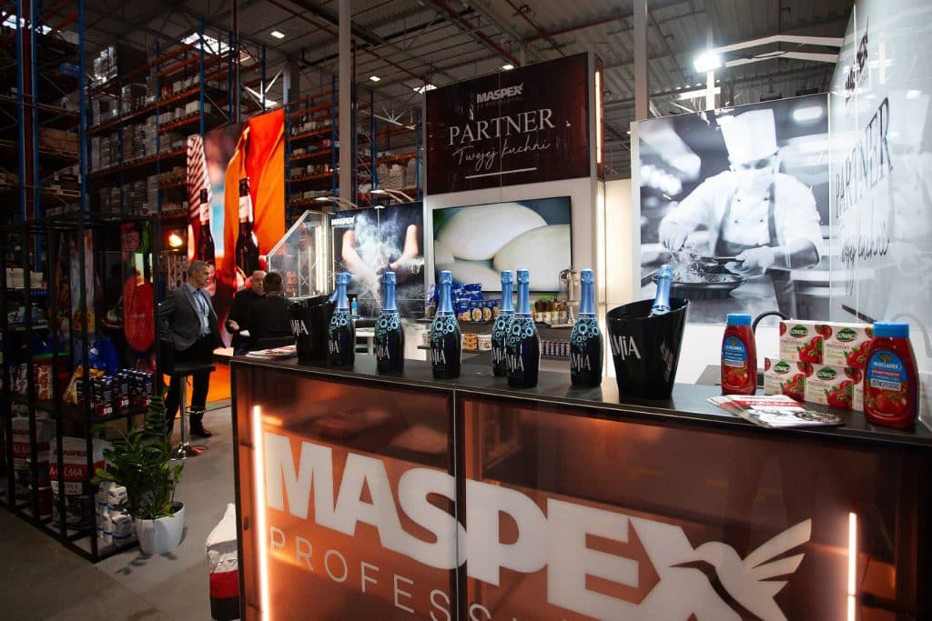 Stoisko z napojami i produktami spożywczymi na targach w Gdańsku, w tle regały z towarami i rozmowy uczestnik&oacute;w.