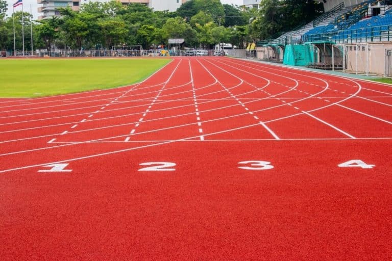 Bieżnia lekkoatletyczna z czerwoną nawierzchnią i białymi liniami startu, na tle zielonej trawy i drzew.
