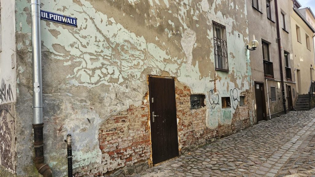 Zniszczona, odrapana ściana budynku z widocznymi cegłami i graffiti na ulicy w Olsztynie.