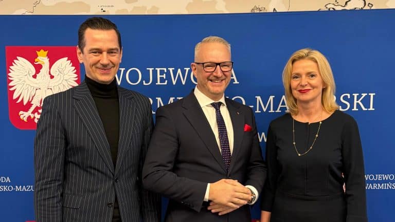Trzy osoby na tle tablicy z napisem 'WOJEWODA WARMIŃSKI' i godłem Polski.