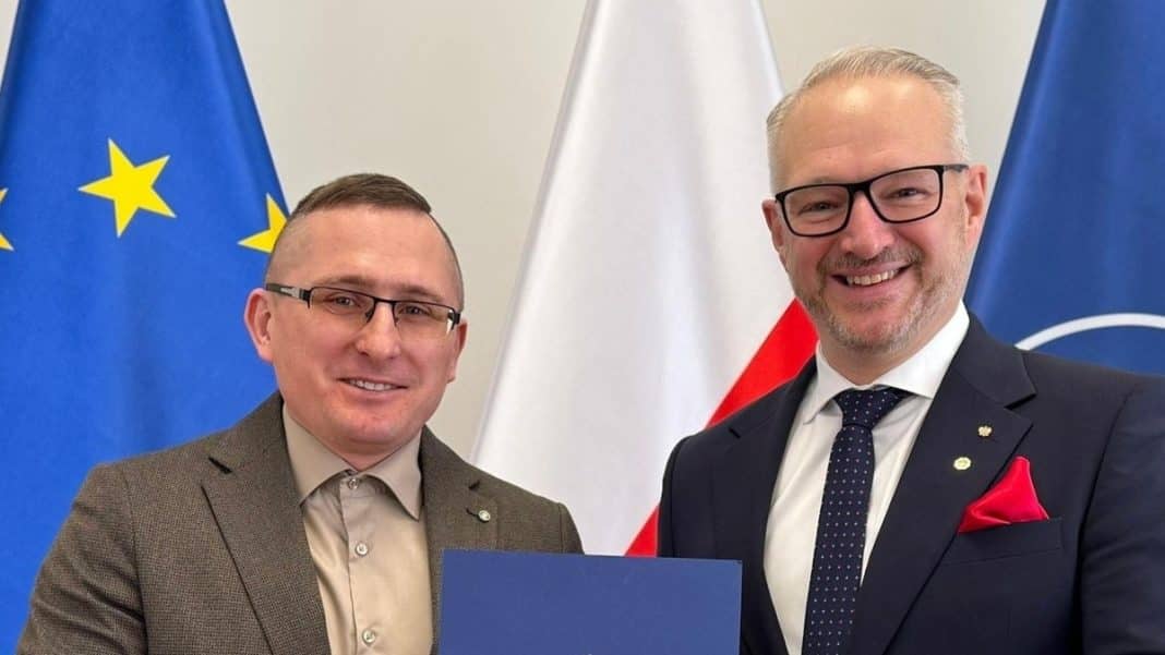 Dw&oacute;ch mężczyzn w garniturach, jeden trzyma dokument, w tle flagi UE i Polski.