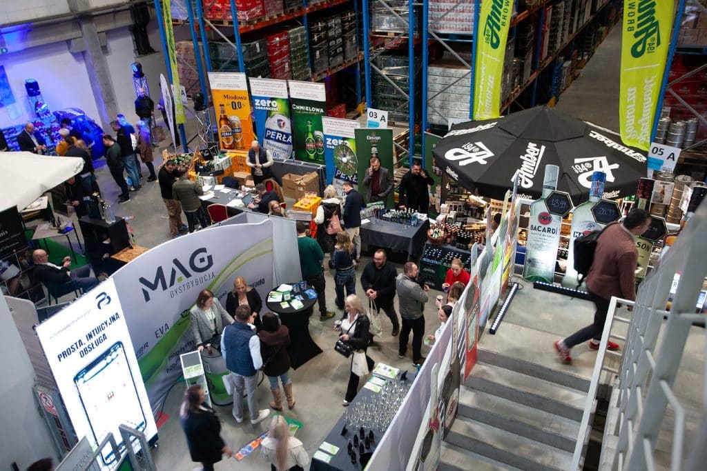 Tłum uczestnik&oacute;w na targach branżowych w hali, stoiska z napojami i produktami, rozmowy i prezentacje.