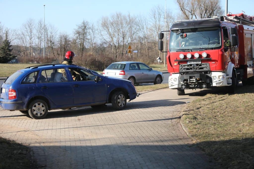 Samoch&oacute;d osobowy dachuje na rondzie w Olsztynie, w pobliżu wozu strażackiego, w tle inne pojazdy i drzewa.