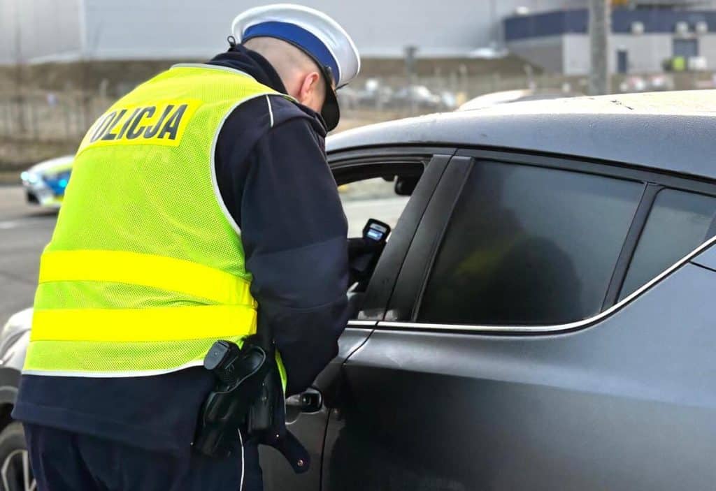 Policjant kontrolujący kierowcę w samochodzie na drodze, w odblaskowej kamizelce.