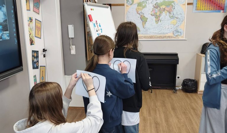 Dzieci w klasie rysują na papierze, stojąc przy tablicy z mapą świata i materiałami edukacyjnymi.