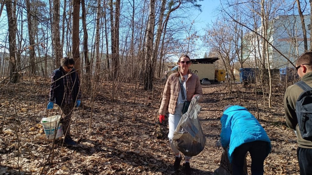 Grupa mieszkańc&oacute;w i wolontariuszy sprząta las w słoneczny dzień, zbierając odpady w okolicy osiedli.