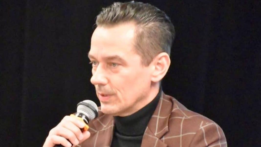 Mężczyzna w brązowej marynarce i czarnym t-shircie, trzymający mikrofon podczas wystąpienia.