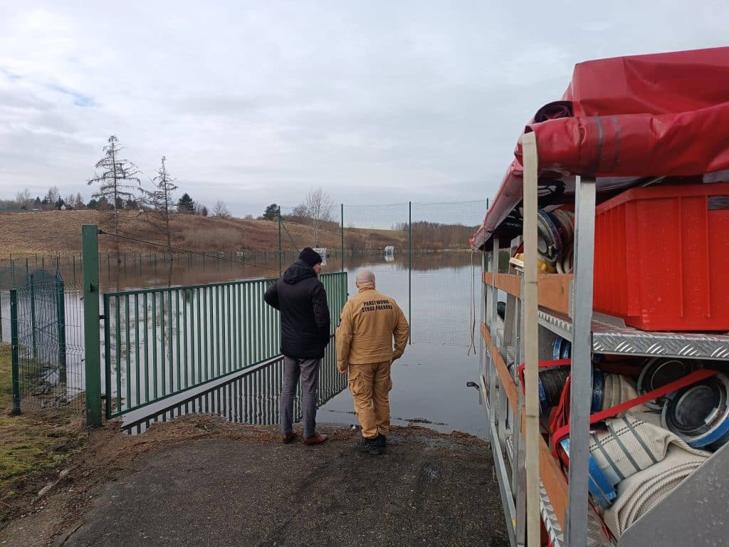 Strażacy i urzędnik obserwują zalane boisko sportowe we Fromborku, w tle woda i pusta przestrzeń.