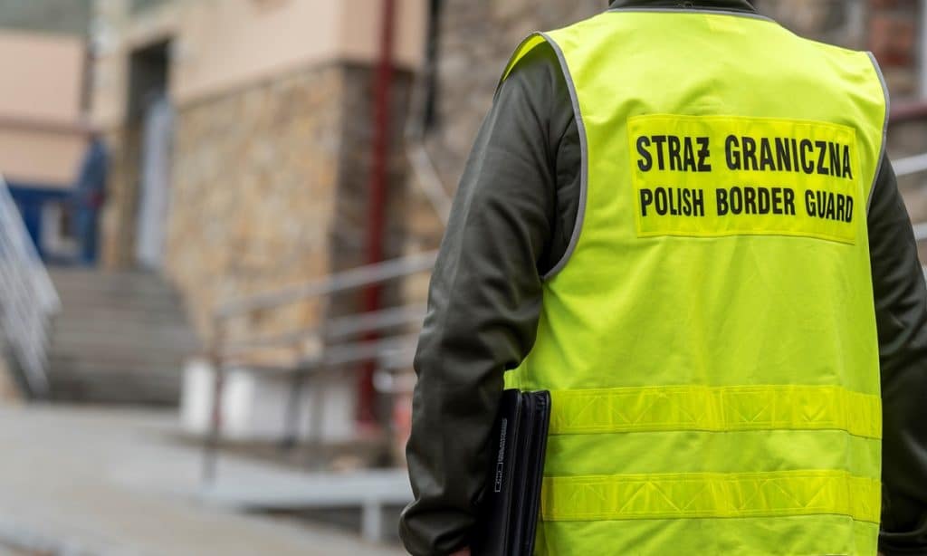 Funkcjonariusz Straży Granicznej w kamizelce odblaskowej na ulicy, z tyłu budynki i barierki.