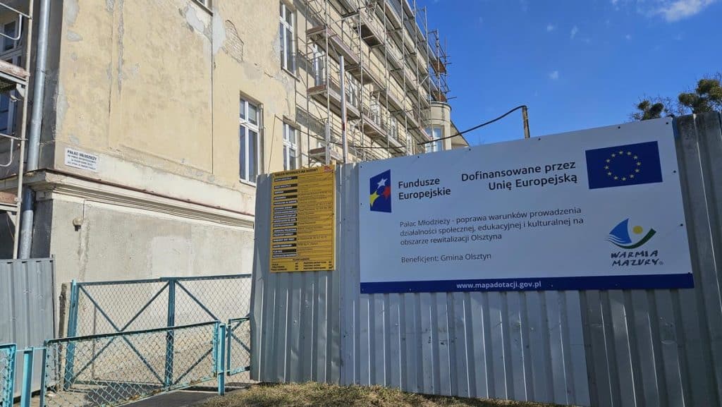 Plac budowy z tablicą informacyjną i rusztowaniami na odrestaurowanym budynku w centrum Olsztyna.