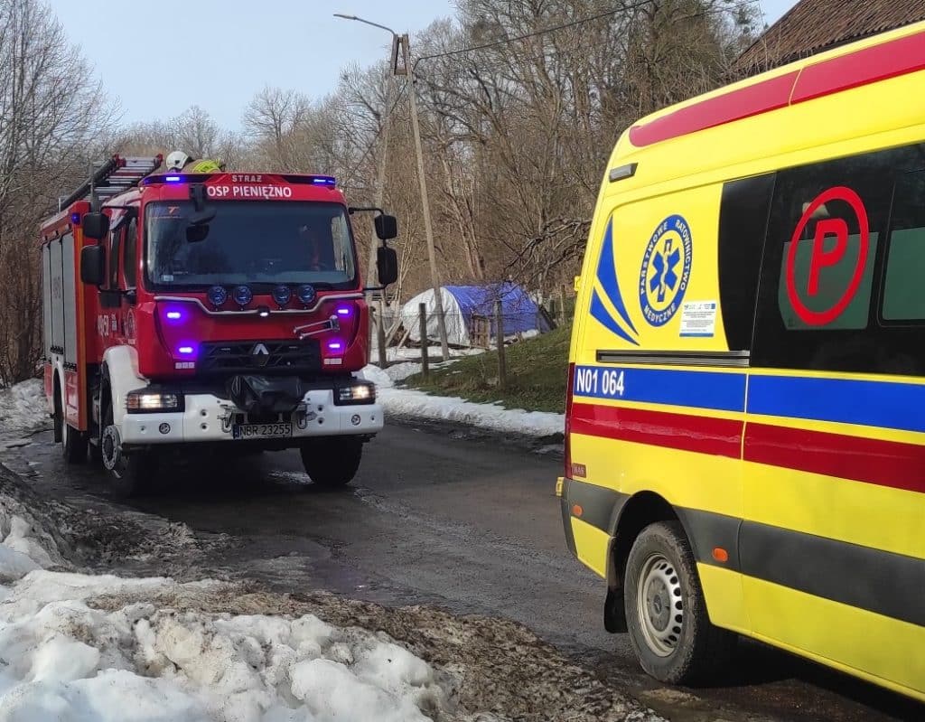 Dwa pojazdy ratunkowe na drodze obok rzeki, w tle drzewa i śnieg, akcja ratunkowa podczas wycinki drzew.
