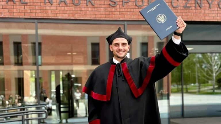 Uczestnik w togach akademickich trzyma dyplom przed budynkiem uczelni.