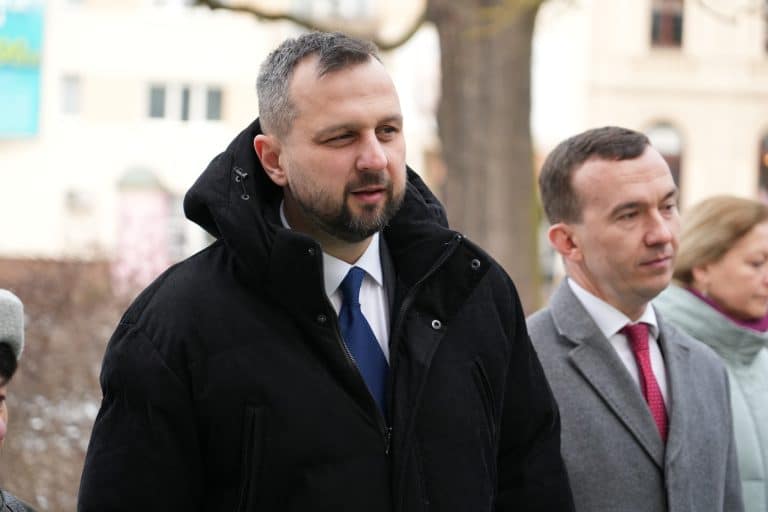 Zdjęcie przedstawia mężczyznę w czarnej kurtce i niebieskiej krawacie, stojącego na zewnątrz, obok innych os&oacute;b.