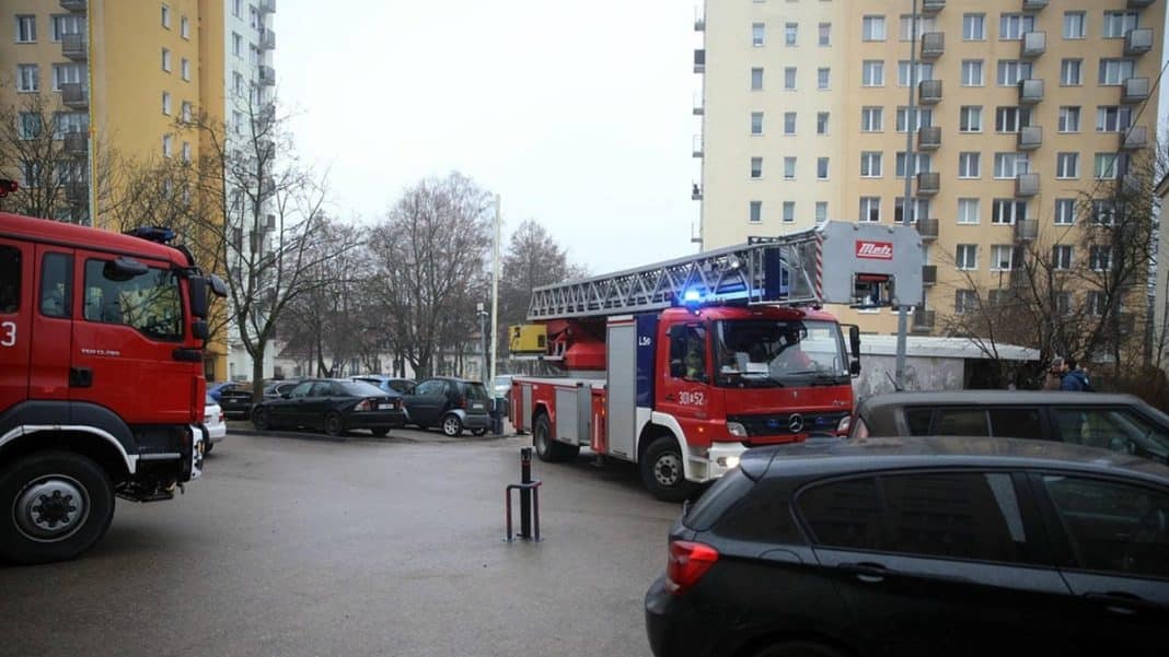 Straż pożarna i służby ratunkowe przed wieżowcem w Olsztynie, w tle bloki mieszkalne.