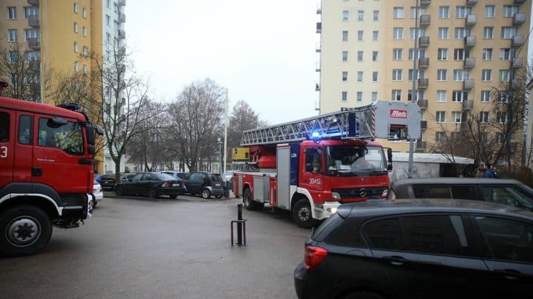 Straż pożarna i służby ratunkowe przed wieżowcem w Olsztynie, w tle bloki mieszkalne.