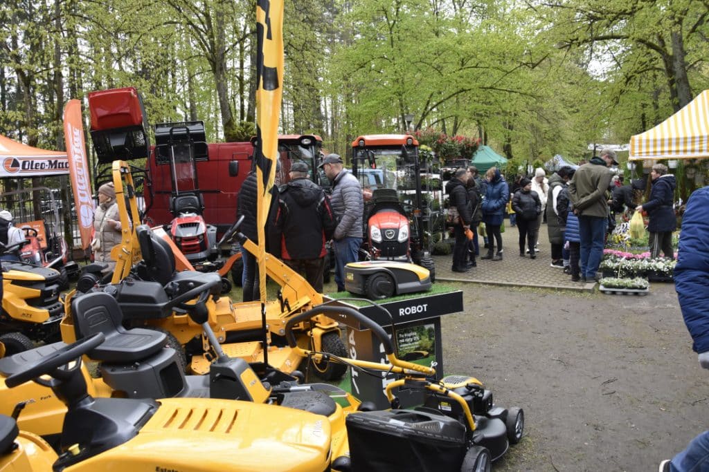Tłum ludzi na targach ogrodniczych z maszynami i roślinami w tle, w parku w Olsztynie.