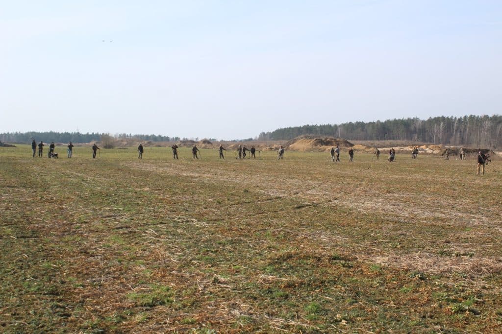 Grupa archeolog&oacute;w na polu podczas wykopalisk w okolicach wsi Leszcz, w tle las i niebo.