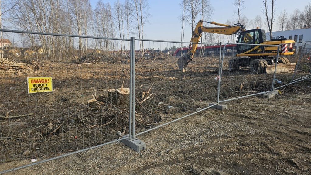 Wycinka drzew na terenie pod budowę nowej siedziby CBZC w Olsztynie, widok na teren z ogrodzeniem i koparką.