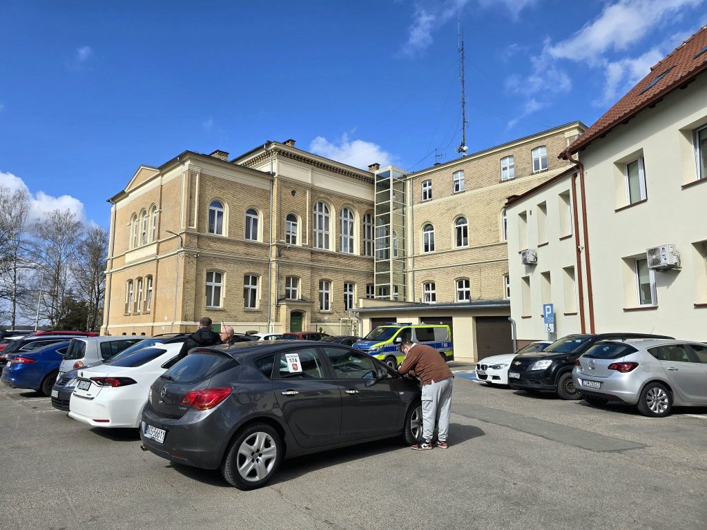 Budynek urzędu miejskiego w Ostr&oacute;dzie, na parkingu przed nim stoją samochody, a na pierwszym planie ludzie rozmawiają przy samochodach.