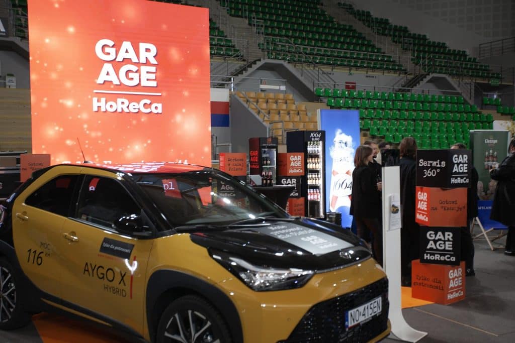 Wewnątrz hali sportowej z ż&oacute;łtym samochodem i stoiskiem targowym, na tle dużego ekranu z napisem GARGE.