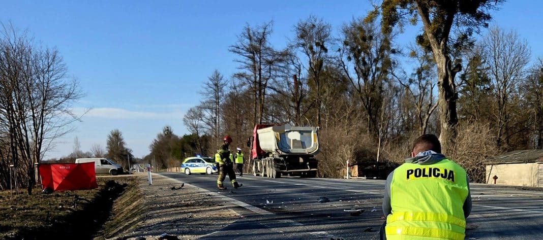 Widok na miejsce wypadku na drodze krajowej z policjantami i służbami ratunkowymi, uszkodzony pojazd ciężarowy.