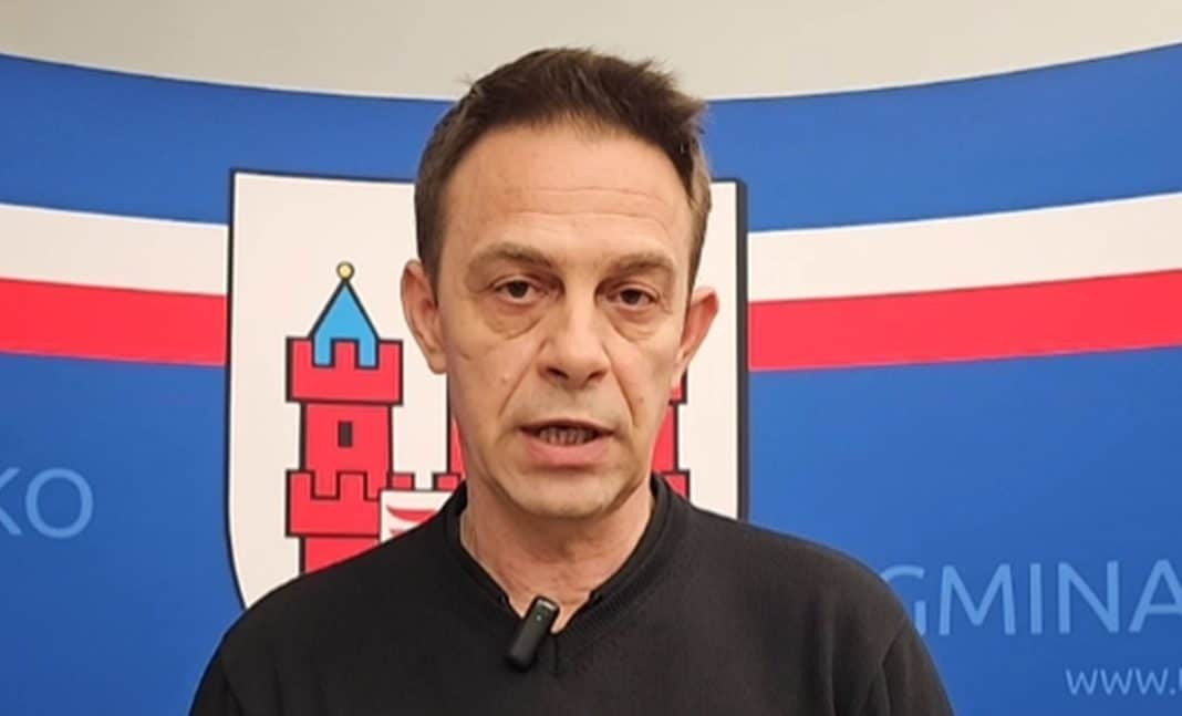 Mężczyzna przemawia przed flagą i herbem gminy na tle niebieskiego tła.