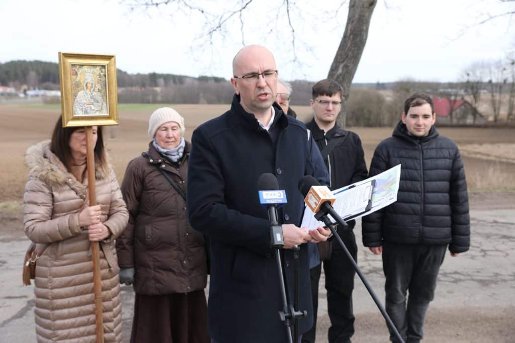 Grupa ludzi na zewnątrz, jedna osoba m&oacute;wi do mikrofonu, w tle pola i drzewa.