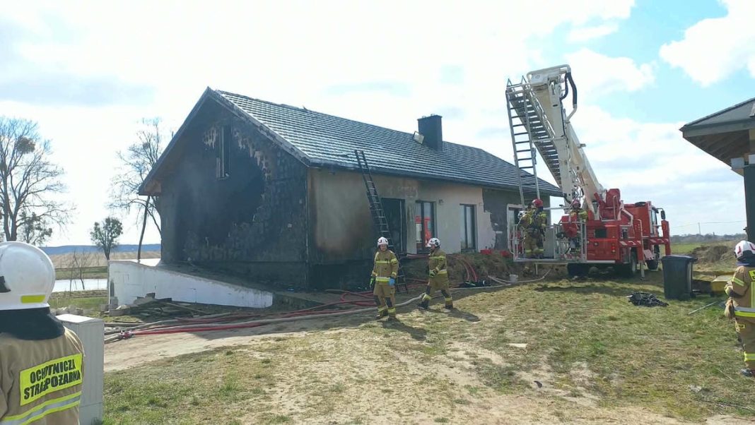 Strażacy gaszą pożar domu jednorodzinnego w Biskupcu, używając drabiny i węży strażackich.