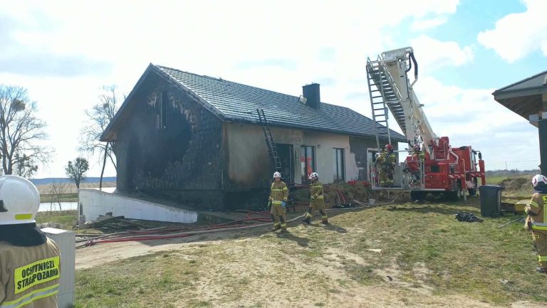 Strażacy gaszą pożar domu jednorodzinnego w Biskupcu, używając drabiny i węży strażackich.