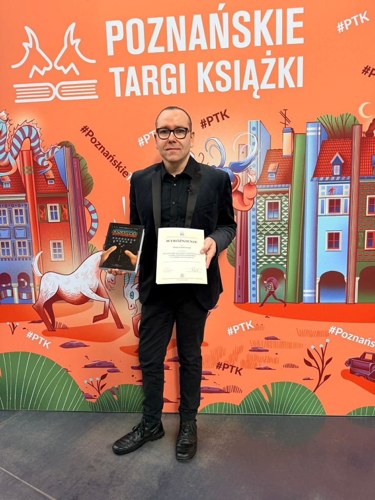 Naukowiec z UWM trzymający nagrodę i certyfikat na tle baneru Targ&oacute;w Książki w Poznaniu.