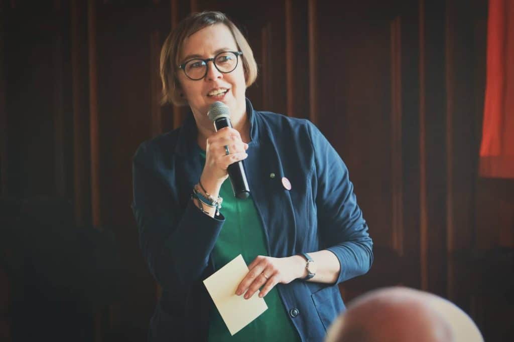 Kobieta z okularami, m&oacute;wiąca do publiczności, trzymająca mikrofon i notatkę, na tle czerwonej kurtyny.