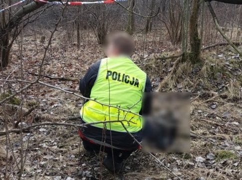 Policjantka bada ciało mężczyzny w lesie, otoczone drzewami i suchą trawą.