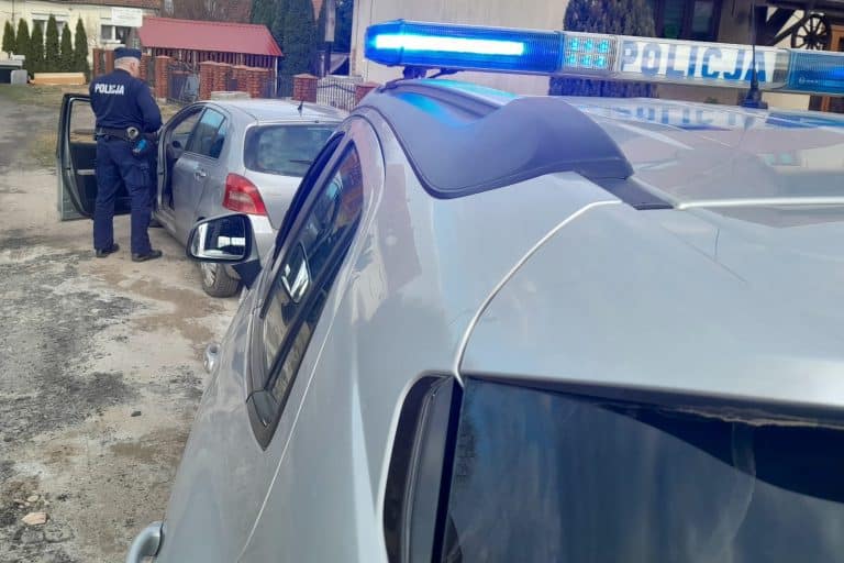 Policjant zatrzymuje samoch&oacute;d na gł&oacute;wnej ulicy, widoczne radiowozy z włączonymi światłami.