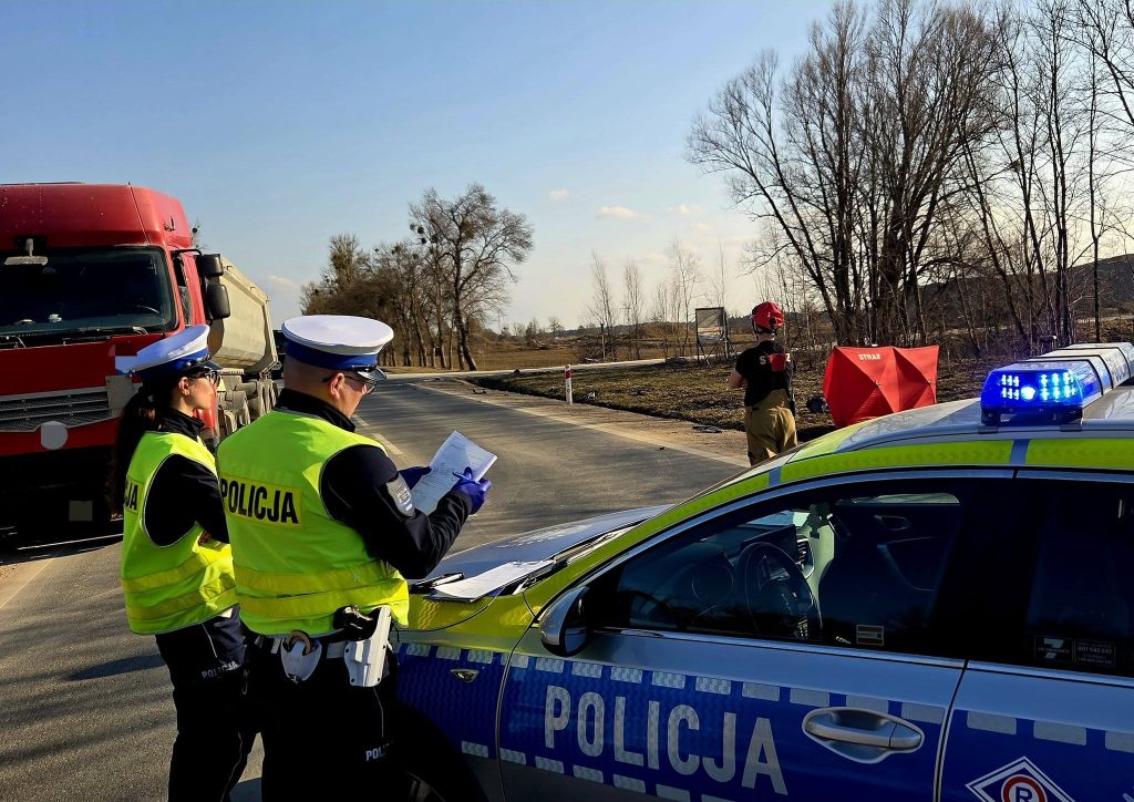 Policjanci i strażacy na miejscu wypadku na drodze krajowej, w tle samoch&oacute;d i ciężar&oacute;wka.
