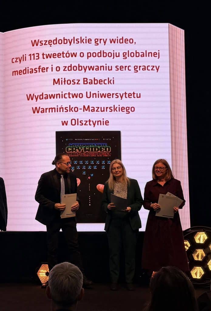 Zdjęcie trzech os&oacute;b na scenie podczas wręczania nagr&oacute;d na targach książki, z dużym ekranem w tle.