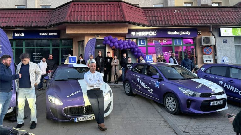 Grupa ludzi i samochody przed nowym punktem szkoły jazdy Hirpol na Jarotach, ozdobionym balonami i plakatami.