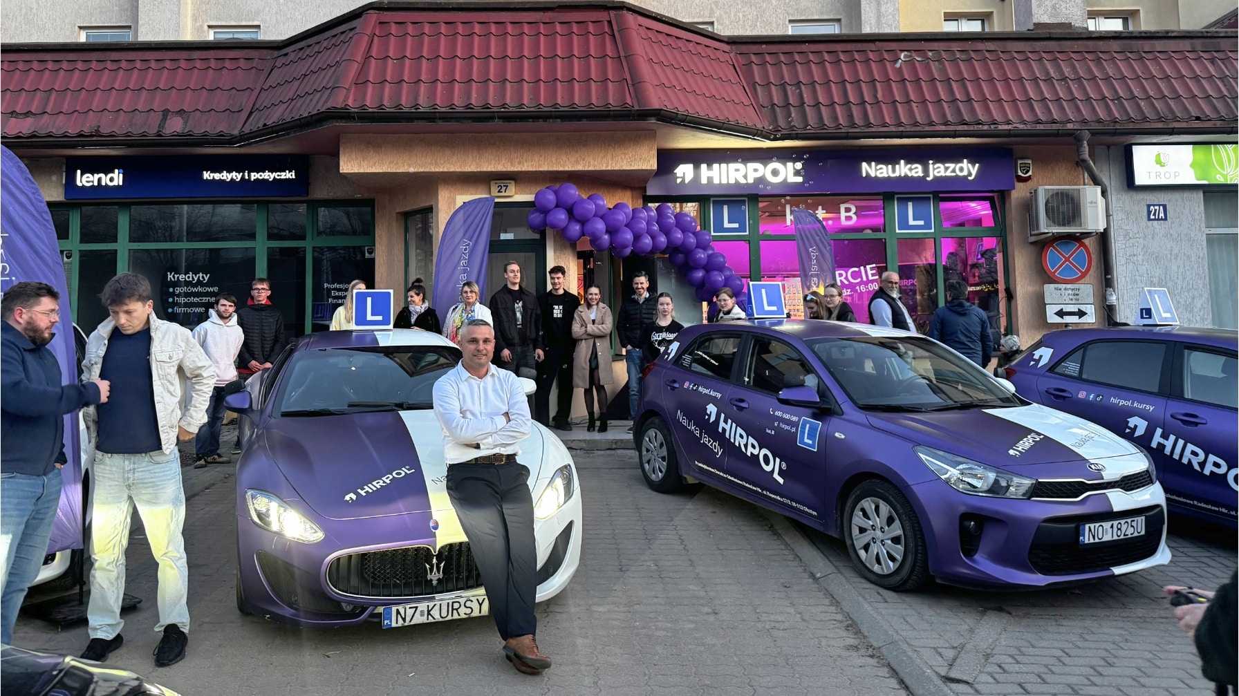 Grupa ludzi i samochody przed nowym punktem szkoły jazdy Hirpol na Jarotach, ozdobionym balonami i plakatami.